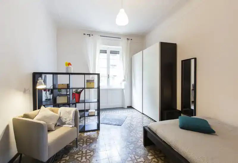 Chambre lumineuse et soignée avec un lit simple, un fauteuil confortable et une étagère ouverte en séparation ; sol carrelé à motifs et grande fenêtre apportant la lumière naturelle.