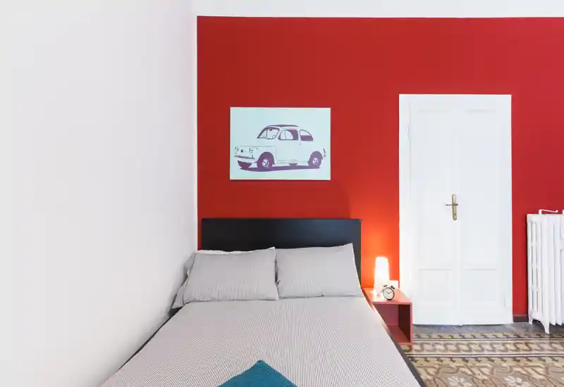 Chambre moderne et lumineuse avec un lit double contre un mur accent rouge, table de chevet avec lampe et une œuvre décorative au-dessus de la tête de lit.