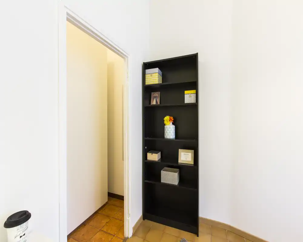 Pequeña entrada o pasillo con una alta estantería negra, suelo de baldosas y una puerta que conduce a una habitación contigua. Paredes blancas y decoración mínima crean un espacio de acceso limpio y sencillo.