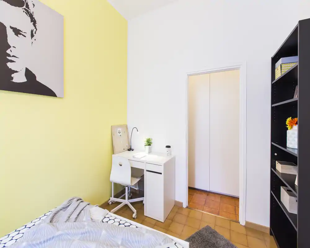 Dormitorio luminoso y ordenado con rincón de cama acogedor, escritorio blanco compacto y estantería — ideal para estudiante o persona sola.