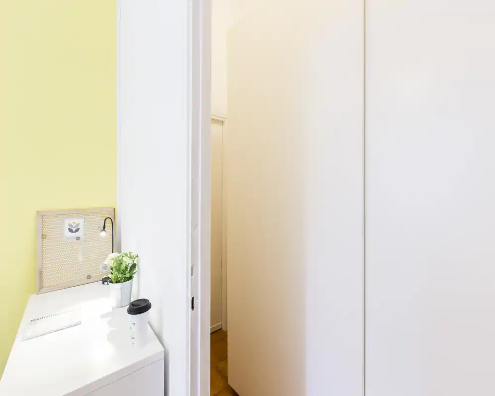 Rincón de trabajo luminoso junto a un pasillo con un pequeño escritorio, planta en maceta y panel de notas: un área ordenada y bien iluminada adecuada para tareas ligeras.