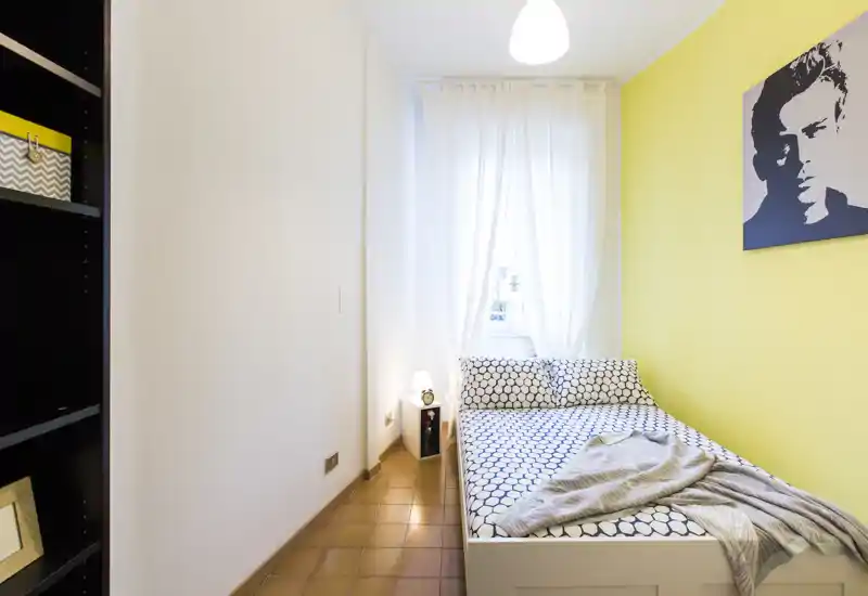 Dormitorio individual luminoso y acogedor con cama de estampado geométrico, ropa de cama suave y lámpara de mesita; ventana alta con cortinas blancas y pared amarilla que da personalidad.