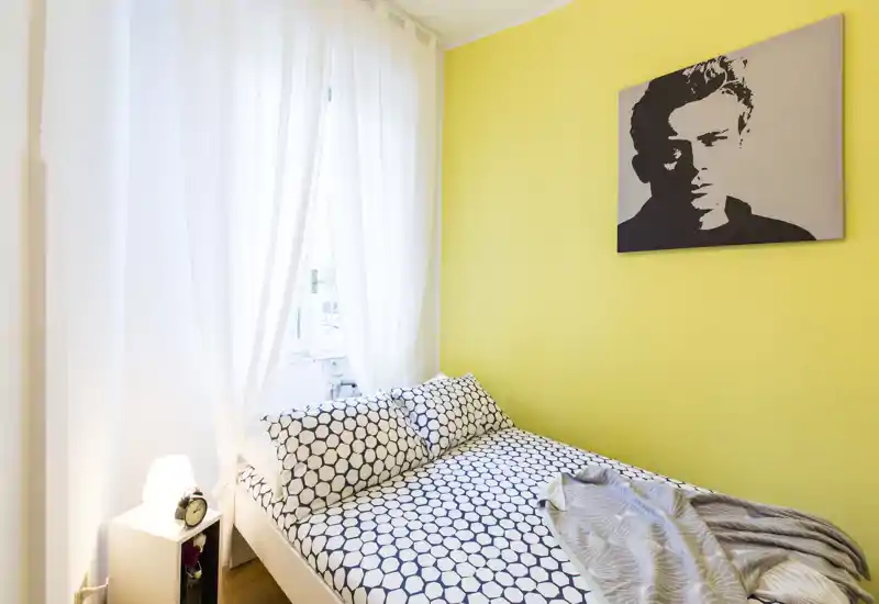 Dormitorio luminoso y compacto con una cama doble estampada frente a una pared amarilla y cortinas ligeras que dejan entrar la luz natural.