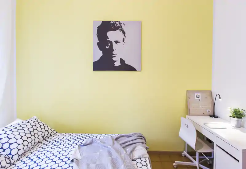Dormitorio luminoso y ordenado con edredón estampado, pequeño espacio de trabajo y retrato sobre pared amarilla — ideal para estudiante o persona sola.