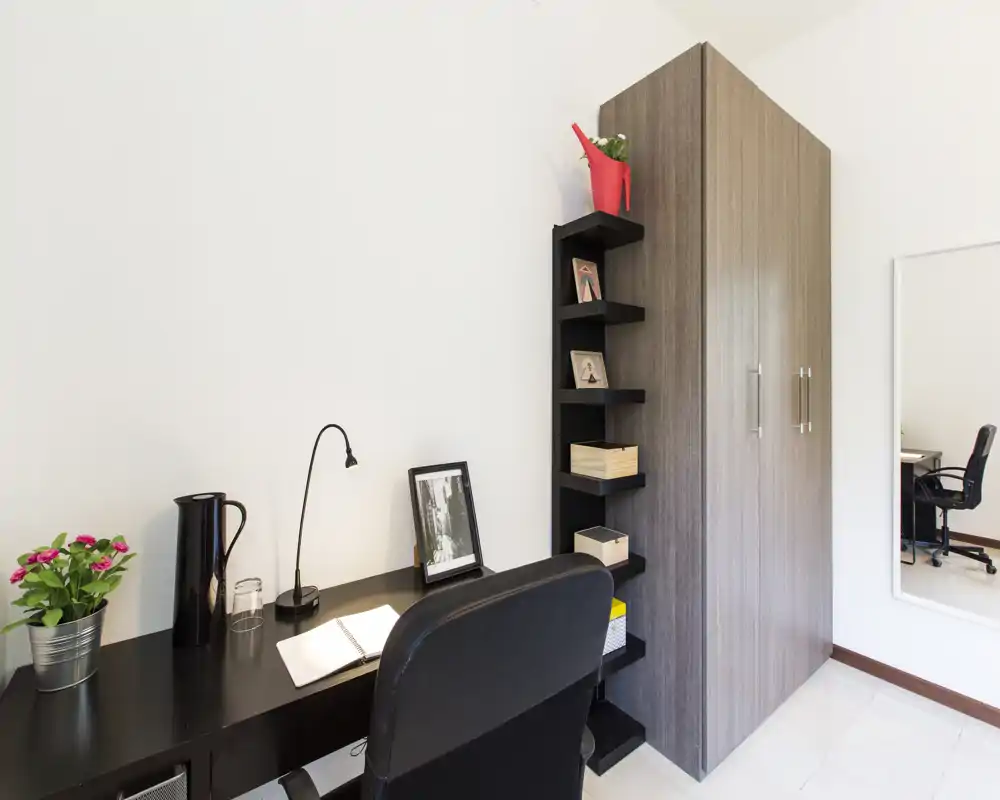 Angolo di lavoro luminoso e minimale con scrivania nera, sedia da ufficio, scaffali e un armadio alto: ambiente ordinato e funzionale per studiare o lavorare.