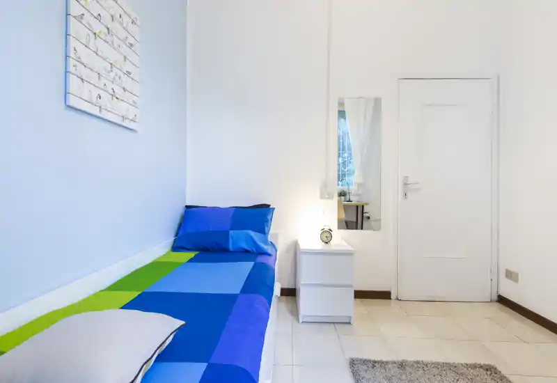 Bright, minimalist single bedroom with a blue-green bedspread, bedside table and lamp; simple, fresh styling ideal for a short-term rental.