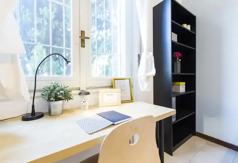 Espacio de trabajo luminoso y bien decorado junto a una gran ventana con escritorio de madera, lámpara, planta y estantería — ideal como oficina en casa o rincón de estudio.