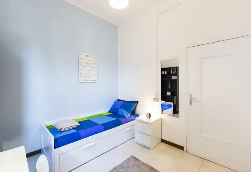 Neat single bedroom with a white daybed, vibrant blue-and-green bedding, bedside table and warm, bright lighting — tidy, modern and ready for a guest.