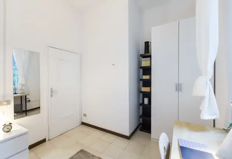 Compact bedroom corner with bright natural light, a wardrobe, shelving and a small desk by the window—clean, minimal and functional.