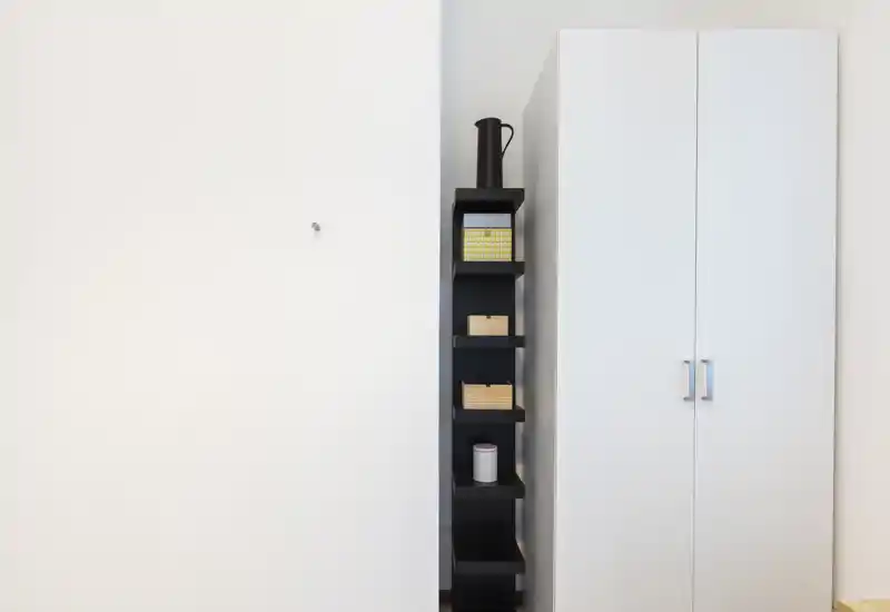 Toma minimalista que muestra una estantería negra estrecha con pequeñas cajas decorativas y un armario blanco de dos puertas contra una pared blanca.