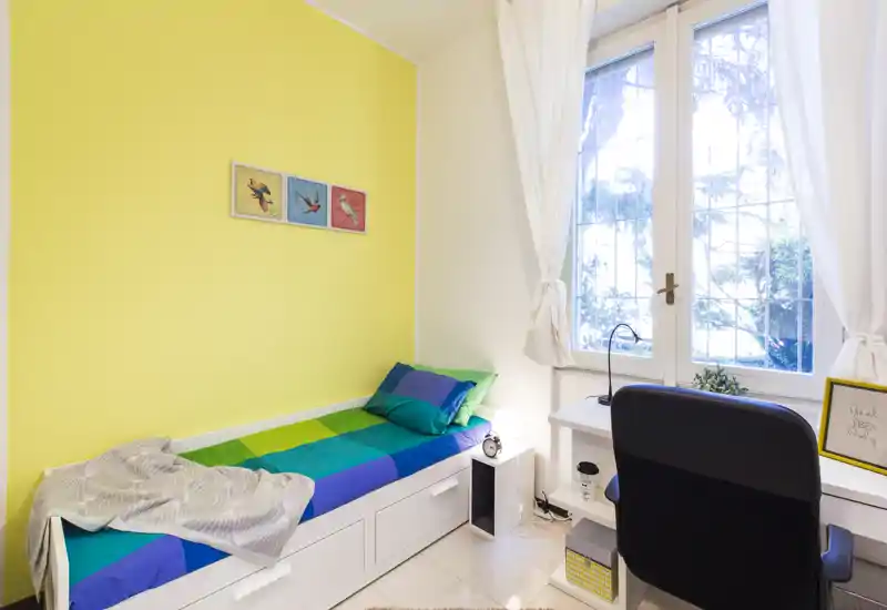 Dormitorio pequeño y luminoso con cama individual de colores y un escritorio junto a una gran ventana que aporta mucha luz natural.