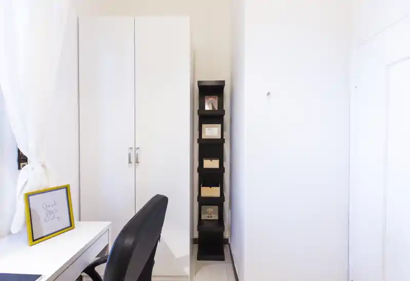 Esquina de trabajo luminosa y minimalista con armario blanco, estantería estrecha y escritorio junto a la ventana con cortina — ordenada y moderna.