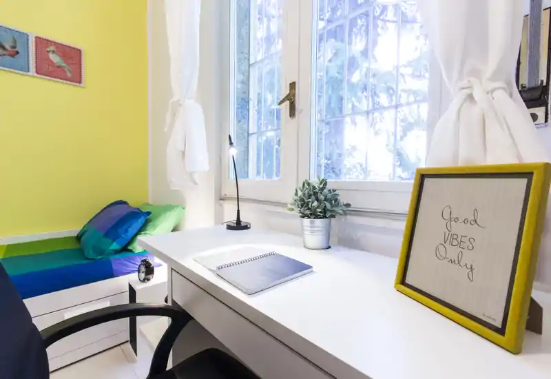 Dormitorio pequeño y luminoso con cama individual, escritorio junto a una gran ventana y decoración alegre — ideal para estudiante o zona de teletrabajo.