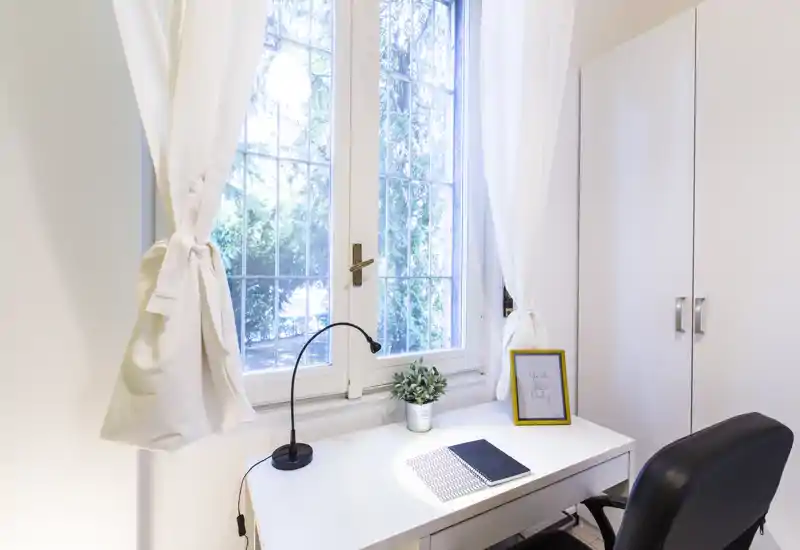 Espacio de trabajo luminoso y ordenado junto a una gran ventana con escritorio blanco, lámpara, planta y silla cómoda — ideal para teletrabajo o estudio.