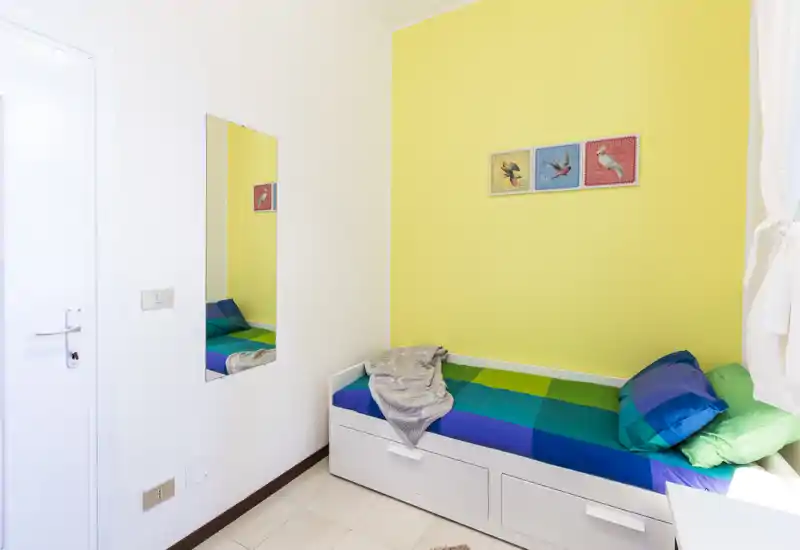 Pequeña habitación individual luminosa con cama colorida frente a una pared amarilla, cajones de almacenaje bajo la cama y luz natural por la ventana.