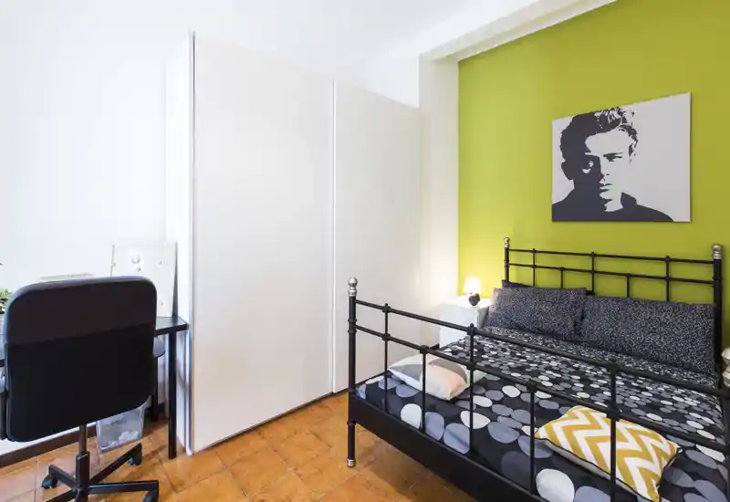 Dormitorio luminoso y ordenado con cama de metal negra frente a una pared verde, ropa de cama con estampado y una pequeña lampara de noche; se aprecia escritorio y armario, ideal también para trabajar.