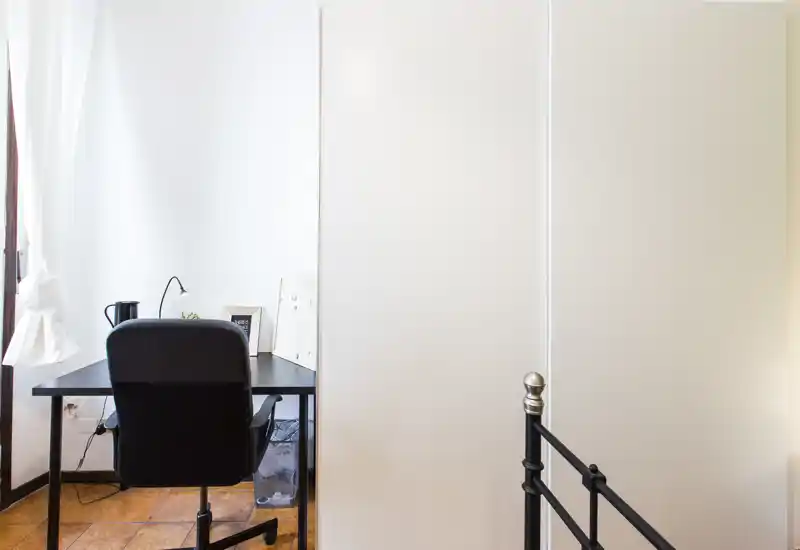 Rincón de dormitorio con la estructura metálica de la cama visible en primer plano a la derecha, un escritorio compacto y silla junto a la ventana y un armario blanco alto que crea un espacio luminoso y minimalista.