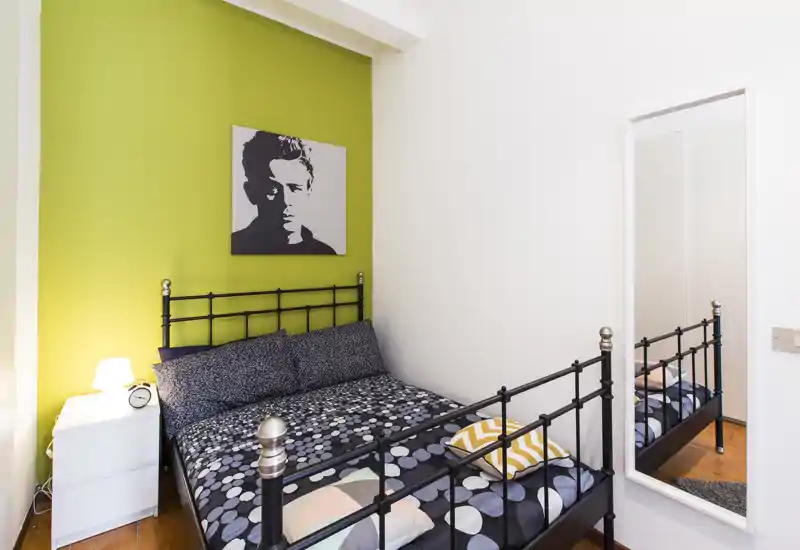 Dormitorio luminoso y con estilo con cama doble de estructura metálica, ropa de cama estampada y pared verde de acento. Mesita con lámpara y espejo de cuerpo entero completan el espacio acogedor y bien iluminado.