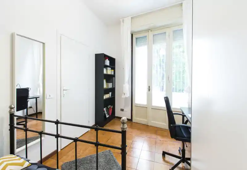 Dormitorio luminoso y ordenado con estructura de cama de metal, escritorio junto a la ventana y estantería — ideal para estudiantes o teletrabajo con buena luz natural.