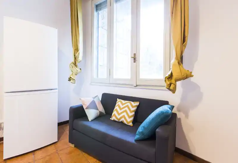 Zona de estar compacta con un sofá oscuro de dos plazas, cojines coloridos y una gran ventana que aporta abundante luz natural—perfecto para un estudio acogedor.