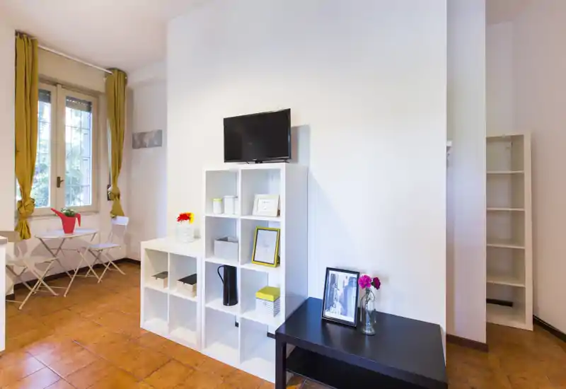 Zona de estar luminosa y bien presentada con estanterías modulares, una pequeña TV y una mesa tipo bistró junto a la ventana — ideal para un apartamento compacto.