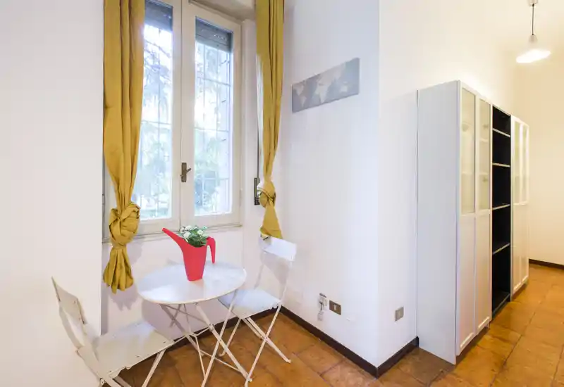 Pequeño rincón con mesa bistró redonda, dos sillas plegables y una ventana con cortinas amarillas; suelo de baldosas y mueble alto a la vista, ideal como estancia o comedor reducido.