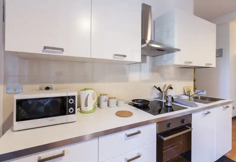 Cocina moderna y luminosa con armarios blancos, placa de inducción, horno empotrado y fregadero doble. Pequeños electrodomésticos y encimera ordenada ofrecen una sensación funcional y cuidada.