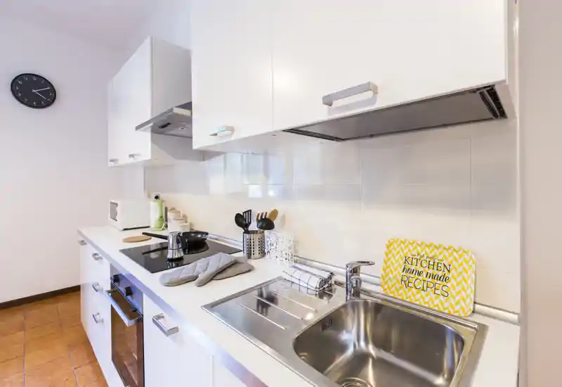 Cocina moderna y luminosa con muebles blancos, fregadero de acero inoxidable, placa de inducción y accesorios en la encimera: espacio limpio y funcional perfecto para estancias cortas.