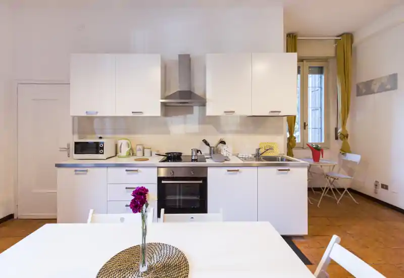 Cocina compacta y moderna con armarios blancos, horno empotrado, campana extractora y encimera ordenada; espacio luminoso con pequeña zona de comedor y detalles decorativos.