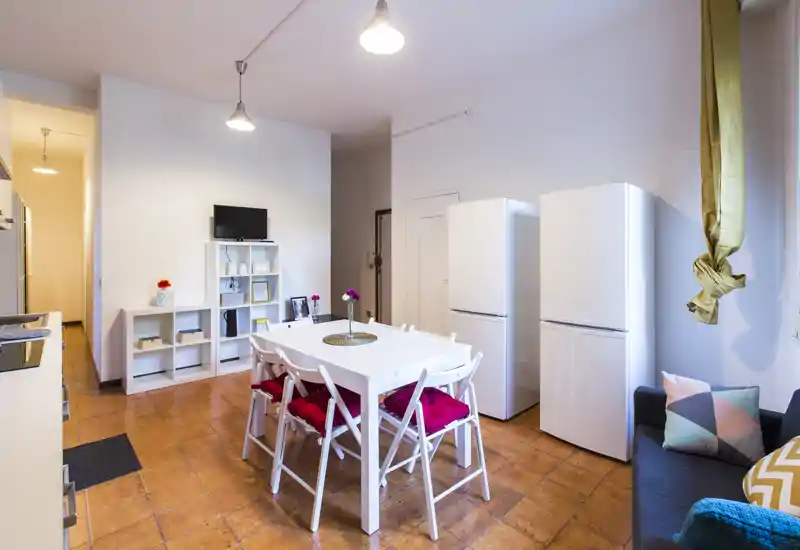 Área de cocina-comedor luminosa con mesa blanca, cuatro sillas con cojines rojos, dos frigoríficos y un pequeño rincón de salón: un espacio práctico y acogedor para comer.