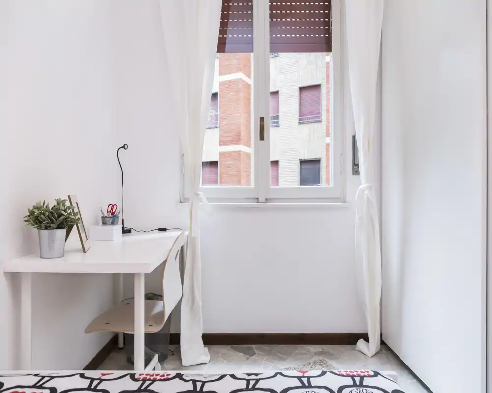 Rincón de dormitorio luminoso y minimalista con un pequeño escritorio junto a la ventana, cortinas claras y el borde de la cama con estampado en primer plano.