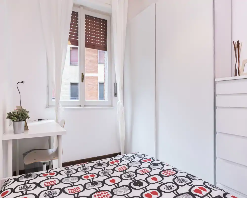 Dormitorio compacto y luminoso con colcha estampada, armario y cómoda blancos; un pequeño escritorio junto a la ventana aporta abundante luz natural.