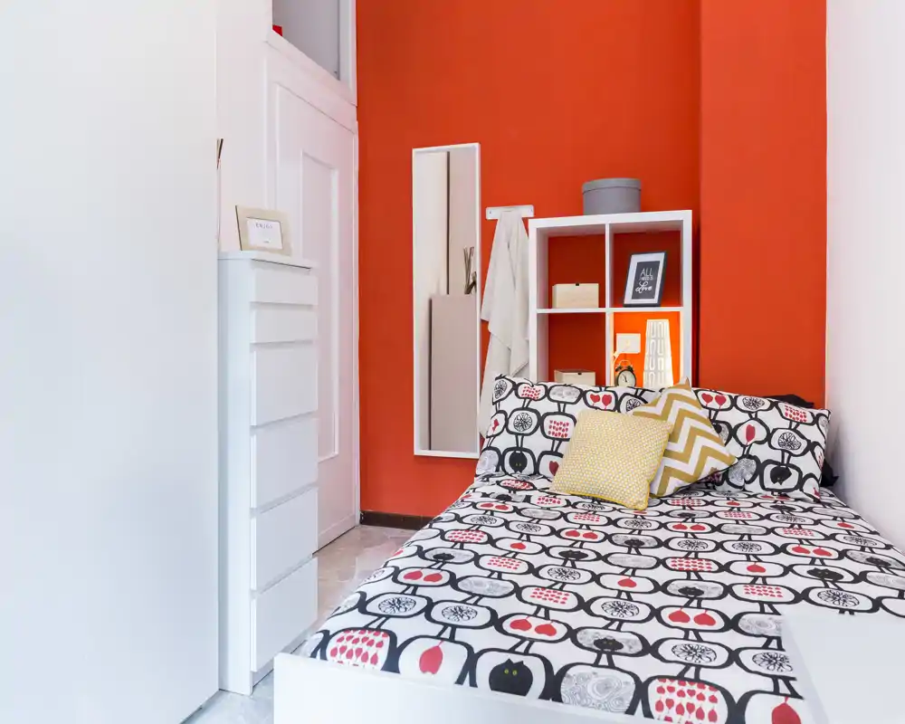 Dormitorio moderno y luminoso con ropa de cama estampada, cojines decorativos y una pared roja llamativa; cama grande y almacenamiento compacto dominan la habitación.
