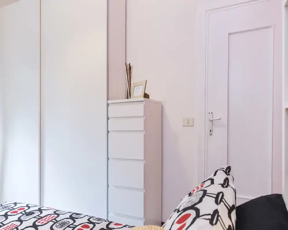 Rincón de dormitorio luminoso y moderno con cama de estampado y una alta cajonera blanca junto a un armario y una puerta cerrada; estilo minimalista y tonos pastel suaves.