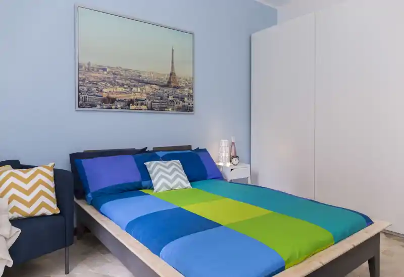 Camera da letto doppia luminosa e ben allestita con piumone a blocchi di colore, cuscini decorativi e una stampa cittadina sopra il letto; comodino con lampada e orologio.