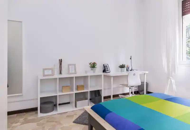 Camera da letto luminosa e moderna con copriletto colorato, scaffalatura minimal e una piccola scrivania vicino alla finestra — ideale per lavoro e relax.