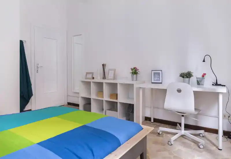 Bright, modern bedroom with a colorful bedcover, white desk and shelving — a clean, well-styled space perfect for rest and study.