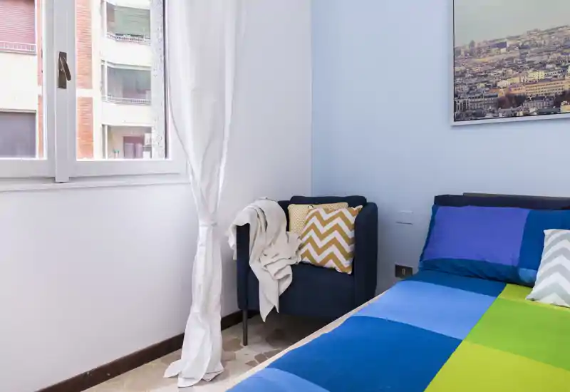 Bright, neatly styled bedroom corner with a colorful bedspread, cushions and a cozy armchair by the window letting in natural light.