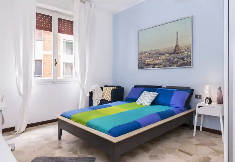 Camera da letto moderna e luminosa con letto ampio con biancheria colorata, comodino con lampada, una poltroncina e una finestra che illumina la stanza.