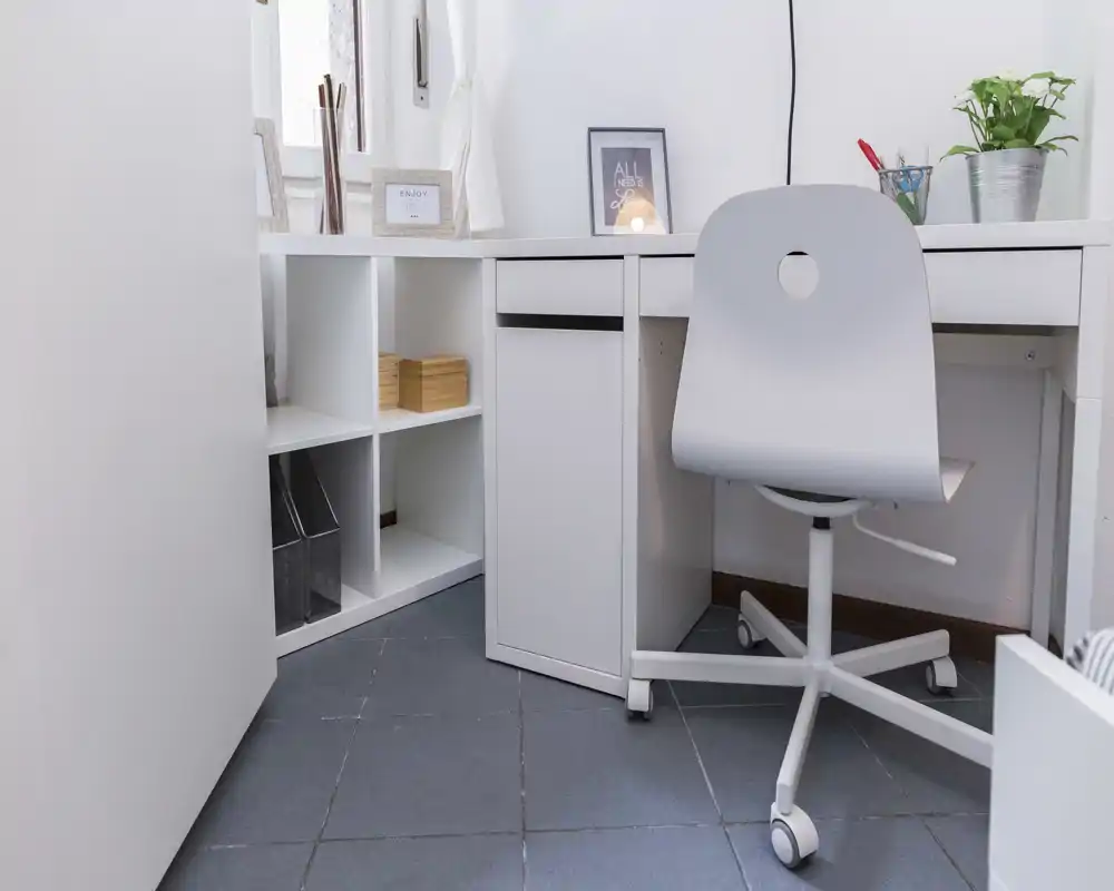Rincón de oficina en casa luminoso y minimalista con escritorio blanco, silla con ruedas y estantería, con una planta pequeña y un cuadro enmarcado.