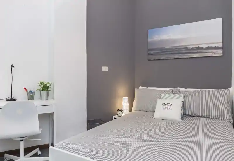 Dormitorio moderno y luminoso con cama doble bien hecha, lámpara de noche y escritorio minimalista — tonos grises y cuadro costero que aportan tranquilidad.