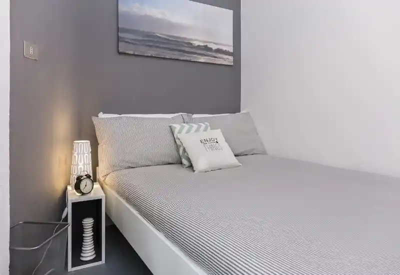 Dormitorio luminoso y ordenado con ropa de cama a rayas, cojines decorativos y lámpara de noche. Un cuadro con un paisaje marino sobre la cama añade un toque moderno y relajante.