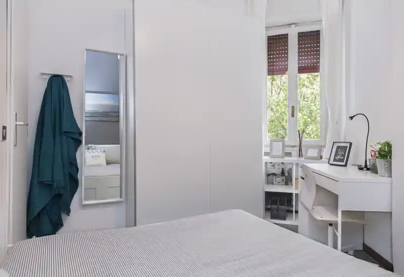Dormitorio luminoso y compacto con cama bien hecha, armario blanco y pequeño escritorio junto a la ventana — ideal para una persona o estancias cortas.