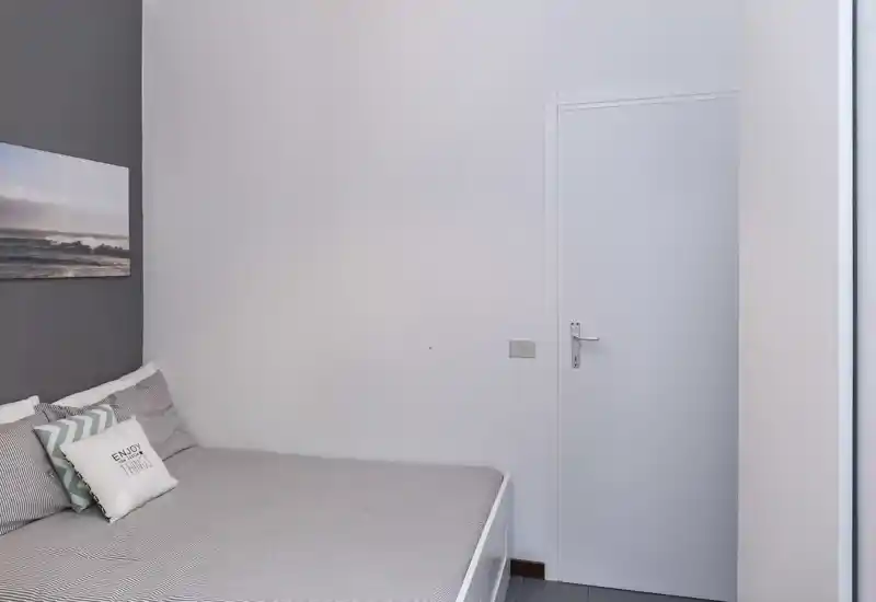 Dormitorio minimalista y bien iluminado con cama ordenada, cojines decorativos y un cuadro costero — tonos neutros y líneas limpias que transmiten tranquilidad.