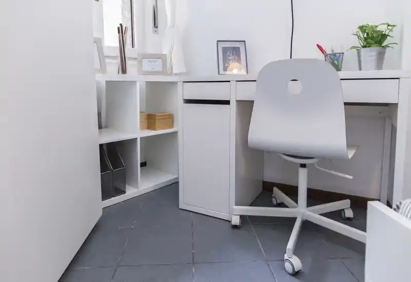 Rincón de oficina en casa luminoso y minimalista con escritorio blanco, silla con ruedas y estantería, con una planta pequeña y un cuadro enmarcado.