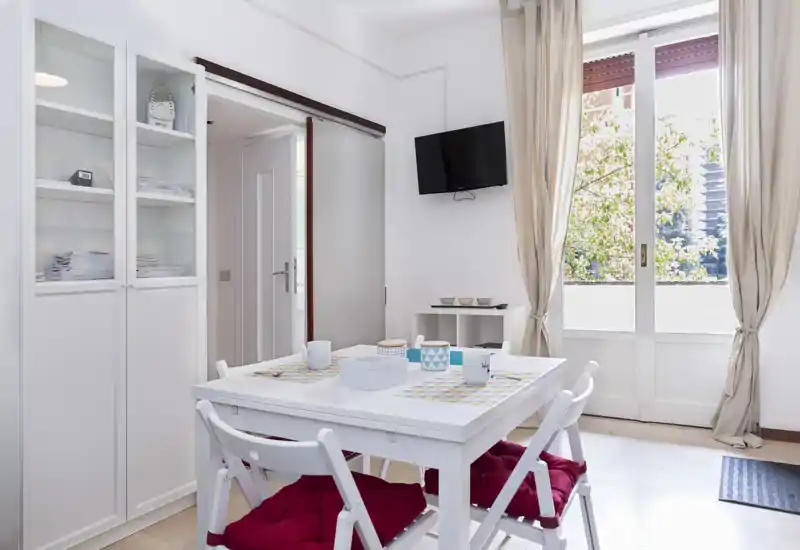 Bright and tidy dining area with a white table, four chairs with red cushions, decorative placemats and a large window providing natural light.