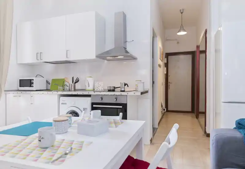 Cocina abierta y luminosa con una pequeña mesa de comedor preparada. Muebles blancos, campana extractora moderna y electrodomésticos compactos crean un espacio funcional ideal para un piso pequeño.