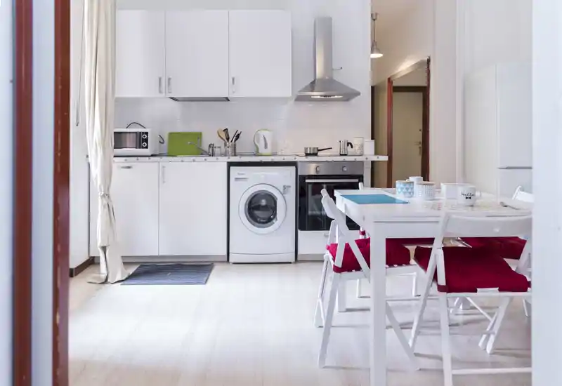 Bright, compact kitchen with white cabinetry, integrated oven, washing machine, and a small dining table with red cushions — ideal for a cozy studio apartment.