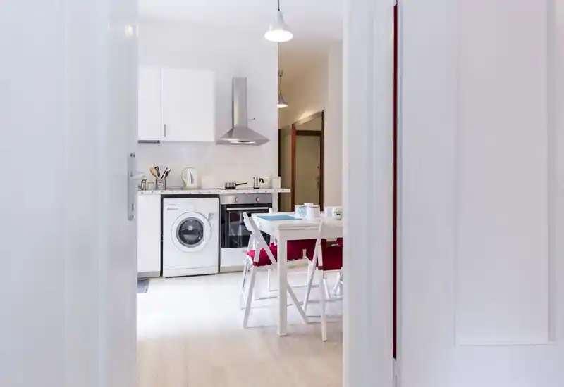 Modern compact kitchen with white cabinets, built-in oven and washing machine, and a small dining table with chairs — bright, neat and well-equipped for a short stay.