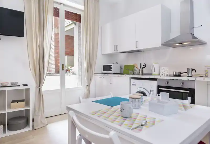 Compact modern kitchen with white cabinets, built-in oven and washing machine, a small dining table set for two and natural light from the glass door leading to a balcony.